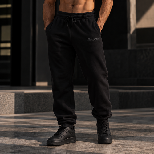  black jogger sweatpants with a small black logo on the left thigh, black sneakers, and a black cap, standing outdoors in front of a modern building.