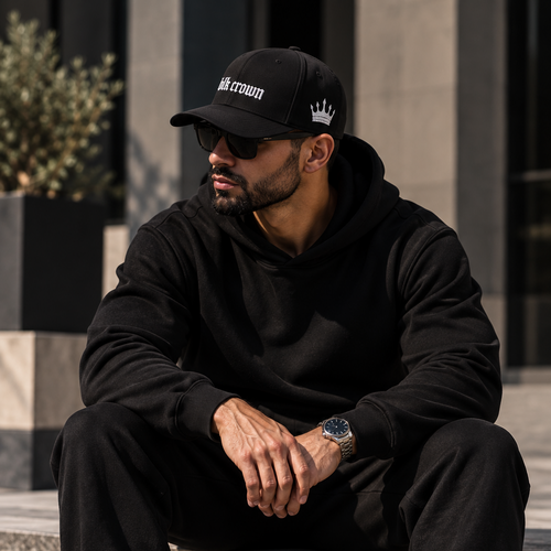 A man wearing a black hoodie, black sweatpants, a black baseball cap with a white crown logo, and white sneakers sits on a stone ledge in an outdoor urban setting.