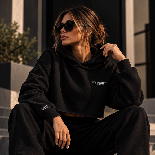 A woman wearing a black cropped hoodie with '666 crown' printed on the chest, black sweatpants, and white sneakers, sitting on outdoor stone steps.
