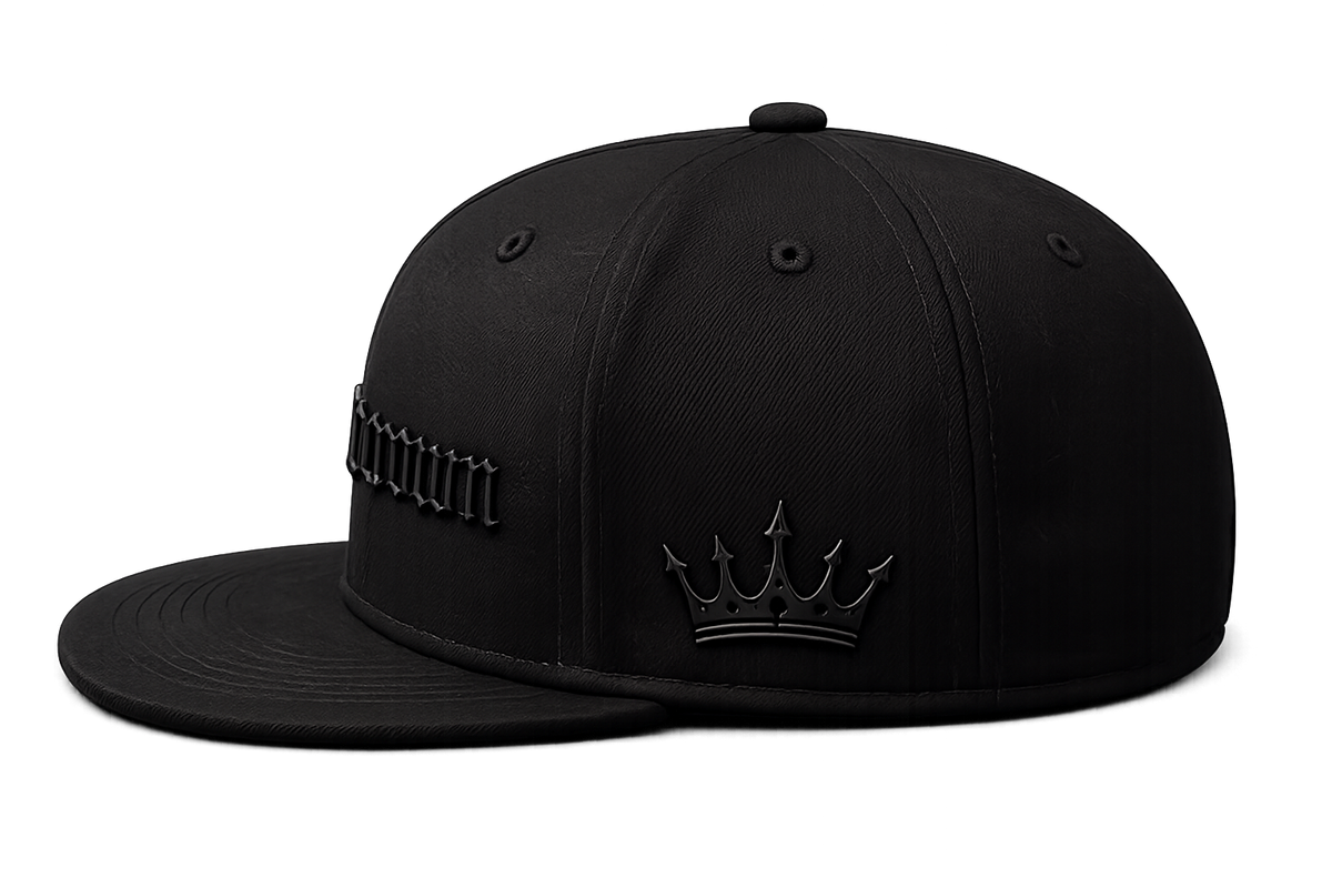 A black structured baseball cap with a flat brim, featuring a tonal black crown design with a crown emblem on the front and stylized text on the side, photographed on a solid white background.
